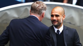 Two football managers facing each other, one with blurred face in formal attire seems to be Pep Guardiola, talking to opponent manager likely during a match setting.