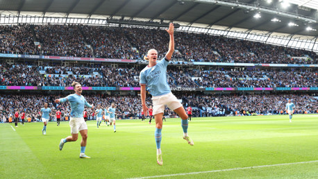 Manchester City player in blue jersey celebrates a goal at Etihad Stadium with enthusiastic crowd in the background.