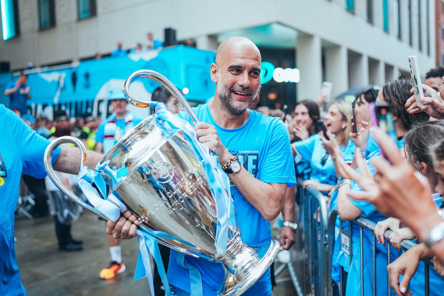 A person in a blue shirt holding a large trophy surrounded by fans. The setting is outdoors with a bus in the background.