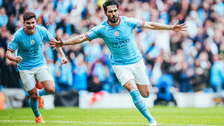 Two Manchester City players celebrating a goal during a match, with one player extending his arms and wearing a captain's armband. They are on the pitch with a stadium crowd in the background.