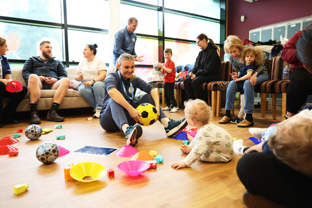 A group of adults and children in a room engaged in play, with toys and a football on the floor. One adult is holding a yellow and black football, and a child is sitting nearby amid colored cones and blocks.