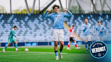 A football player wearing a Manchester City jersey celebrates a goal with his arms flexed. Other players and a goalkeeper are visible on the pitch with empty stands in the background. An 'Official Verdict' badge is in the bottom right corner.