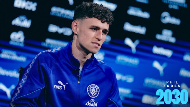 A man in a blue Manchester City tracksuit, possibly an interview setting, with a blurred background featuring logos and text reading 'Phil Foden 2030'.
