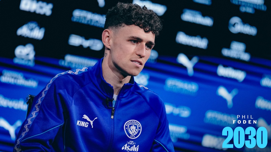 A man in a blue Manchester City tracksuit, possibly an interview setting, with a blurred background featuring logos and text reading 'Phil Foden 2030'.
