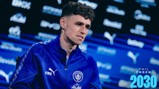 A man in a blue Manchester City tracksuit, possibly an interview setting, with a blurred background featuring logos and text reading 'Phil Foden 2030'.
