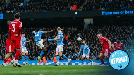Players from Manchester City and another team in red compete for the ball during a match. The focus is on a Manchester City player in light blue attempting a shot or pass.