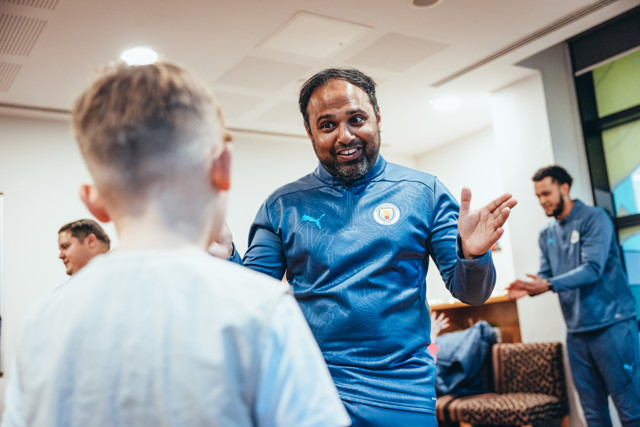 A Manchester City coach, wearing a blue Puma jacket with the team logo, interacts with young player escorts indoors.