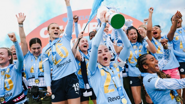 Group of women in light blue jerseys celebrating with a trophy, holding medals and wearing the number 26. They appear joyful and are surrounded by confetti and blue sky.