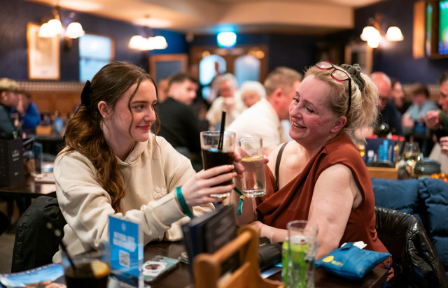 Two women are clinking glasses in a busy pub setting, smiling and enjoying their drinks.