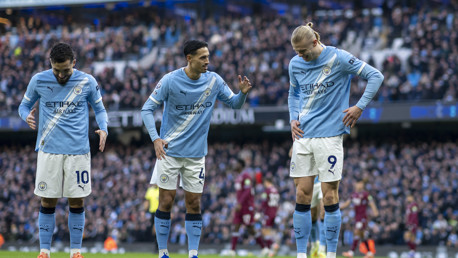 Three Manchester City players in home kit discussing tactics on the field during a match.