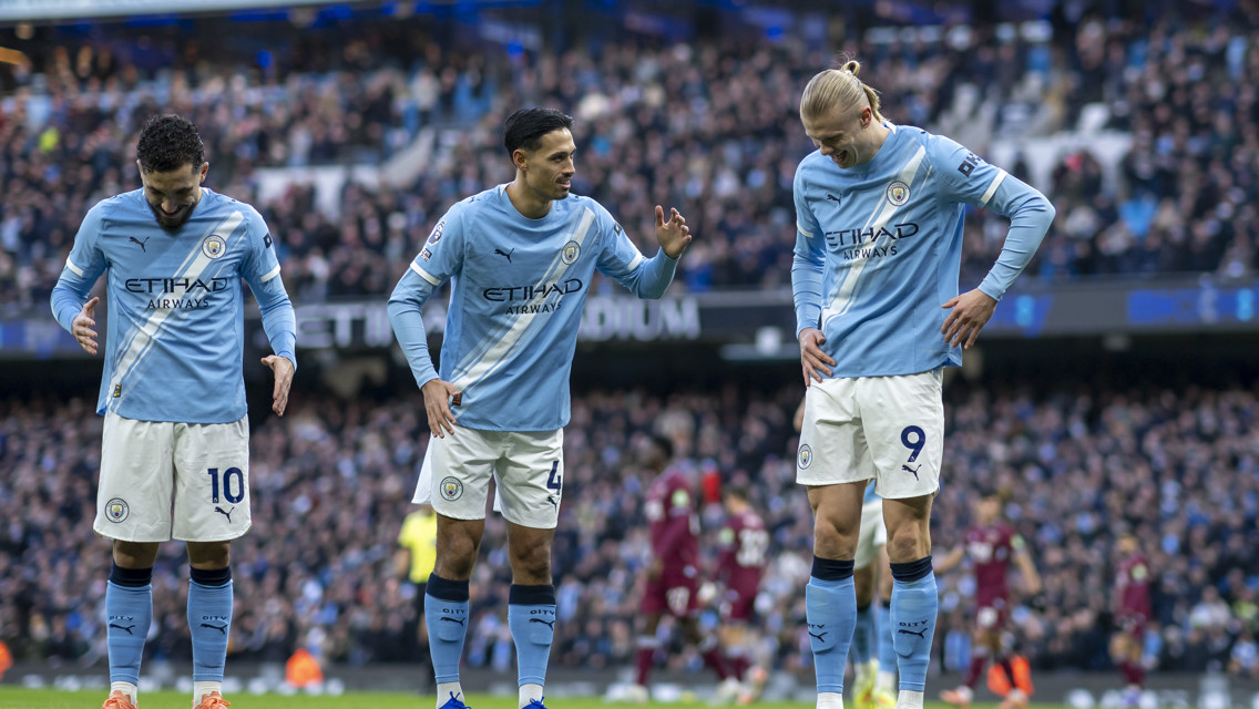 Three Manchester City players in home kit discussing tactics on the field during a match.