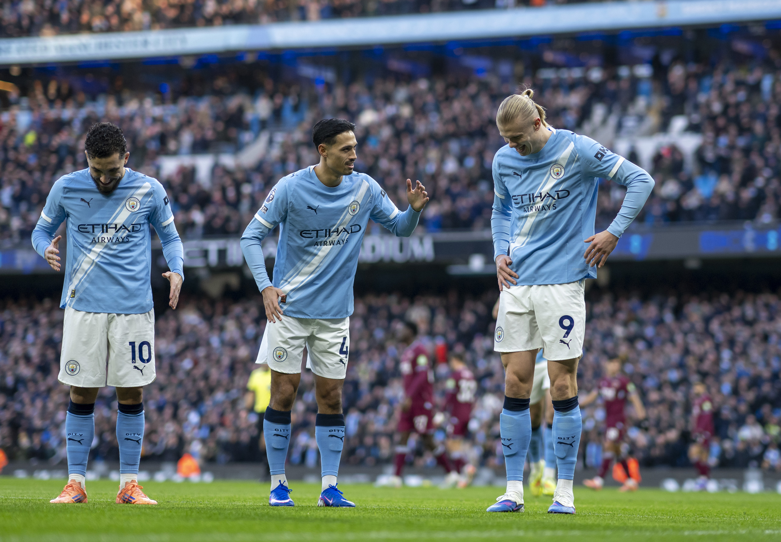 Three Manchester City players in home kit discussing tactics on the field during a match.