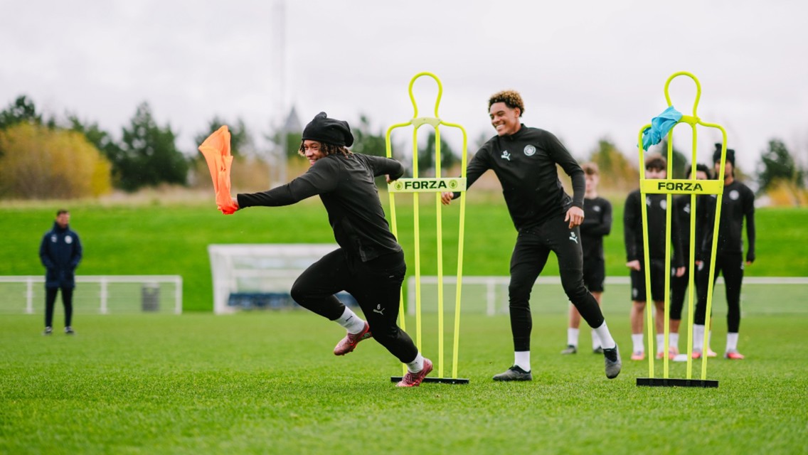 Players in black training gear perform running drills on a football field, maneuvering through mannequins with brightly colored cloth tied around them. One player holds a red cloth in hand running past the mannequin.