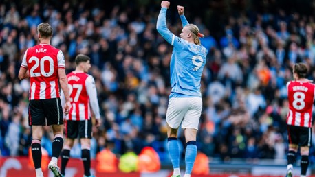 A soccer player in a light blue jersey with the number 9 celebrates a goal by raising their fists. Opponents in red and white jerseys look on.