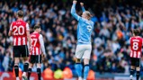 A soccer player in a light blue jersey with the number 9 celebrates a goal by raising their fists. Opponents in red and white jerseys look on.