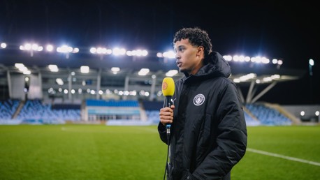 An individual with a blurred face holding a BBC Radio Manchester microphone, standing on a football pitch with a Manchester City logo on his jacket at an empty, illuminated stadium.
