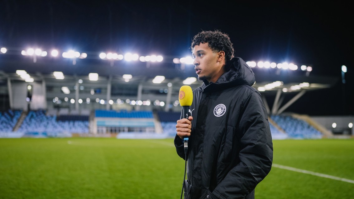 An individual with a blurred face holding a BBC Radio Manchester microphone, standing on a football pitch with a Manchester City logo on his jacket at an empty, illuminated stadium.