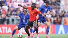 Three soccer players in action during a match. The player in a red jersey is trying to control the ball against two players in blue patterned jerseys.