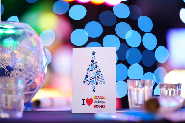 A holiday card featuring a stylized Christmas tree design in blue and white patterns and 'I love 1990s 2000s' text. Surrounded by colored lights and glass cups.