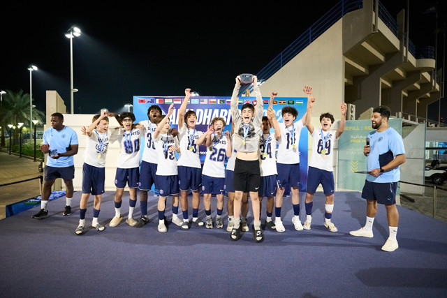 A group of people wearing matching sports uniforms celebrate on a stage. One person holds a trophy above their head. The background includes a banner with international flags and the text 'Abu Dhabi Cup.'