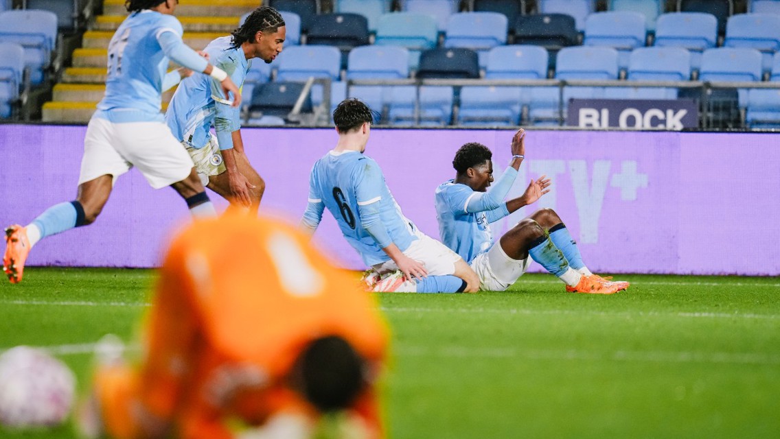 Manchester City players celebrating a goal during a football match, with a goalkeeper in an orange outfit on the ground. The image shows players seated and raising their hands.