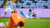 Manchester City players celebrating a goal during a football match, with a goalkeeper in an orange outfit on the ground. The image shows players seated and raising their hands.