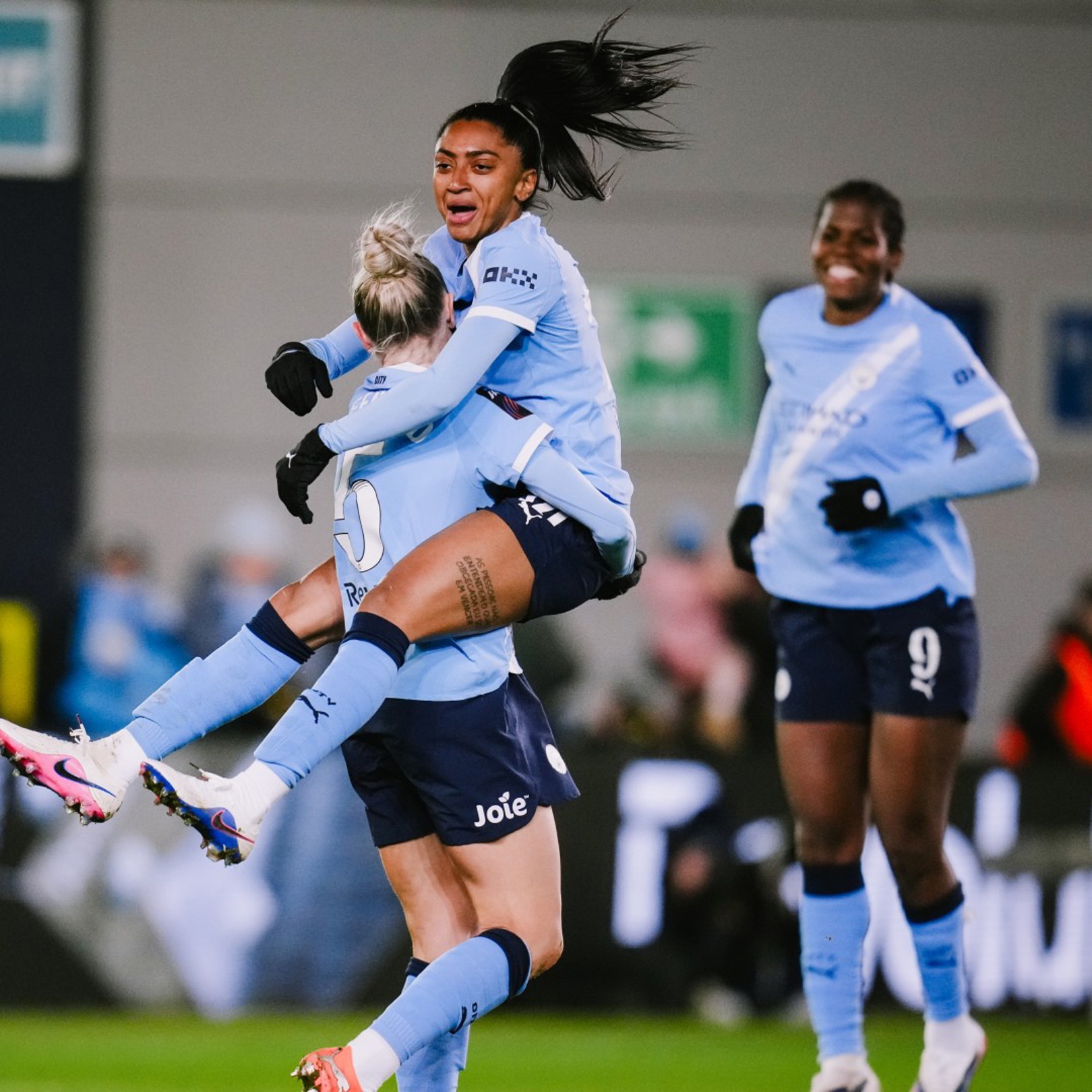 Players wearing light blue and dark blue jerseys celebrate a goal on a football field. One player is lifted in the air by a teammate.