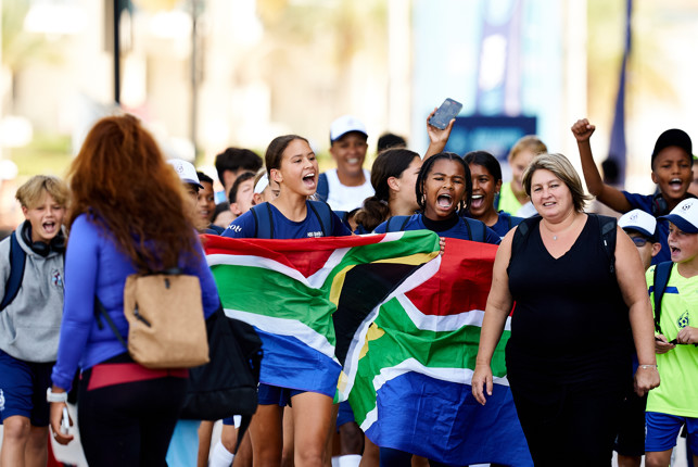 A group of children holding a South African flag, accompanied by two adults, in a public setting.