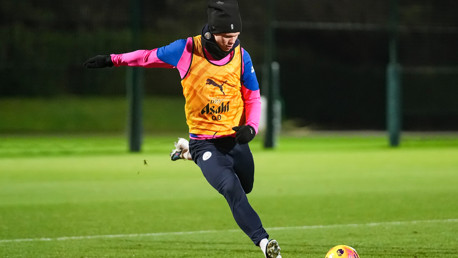 A soccer player is training on a field, kicking a ball. They are wearing a pink and blue jersey with an orange bib that has a Puma logo and 'Asahi' text.