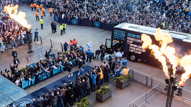 An aerial view of Manchester City team arriving at the Etihad Stadium, greeted by fans and security, with a flame show.
