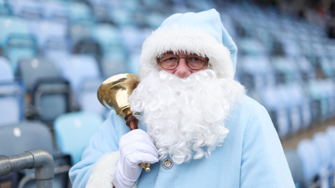 A person dressed as Santa Claus in a blue outfit, holding a bell with a brass top and wooden handle. The scene is set in a stadium with rows of seating.