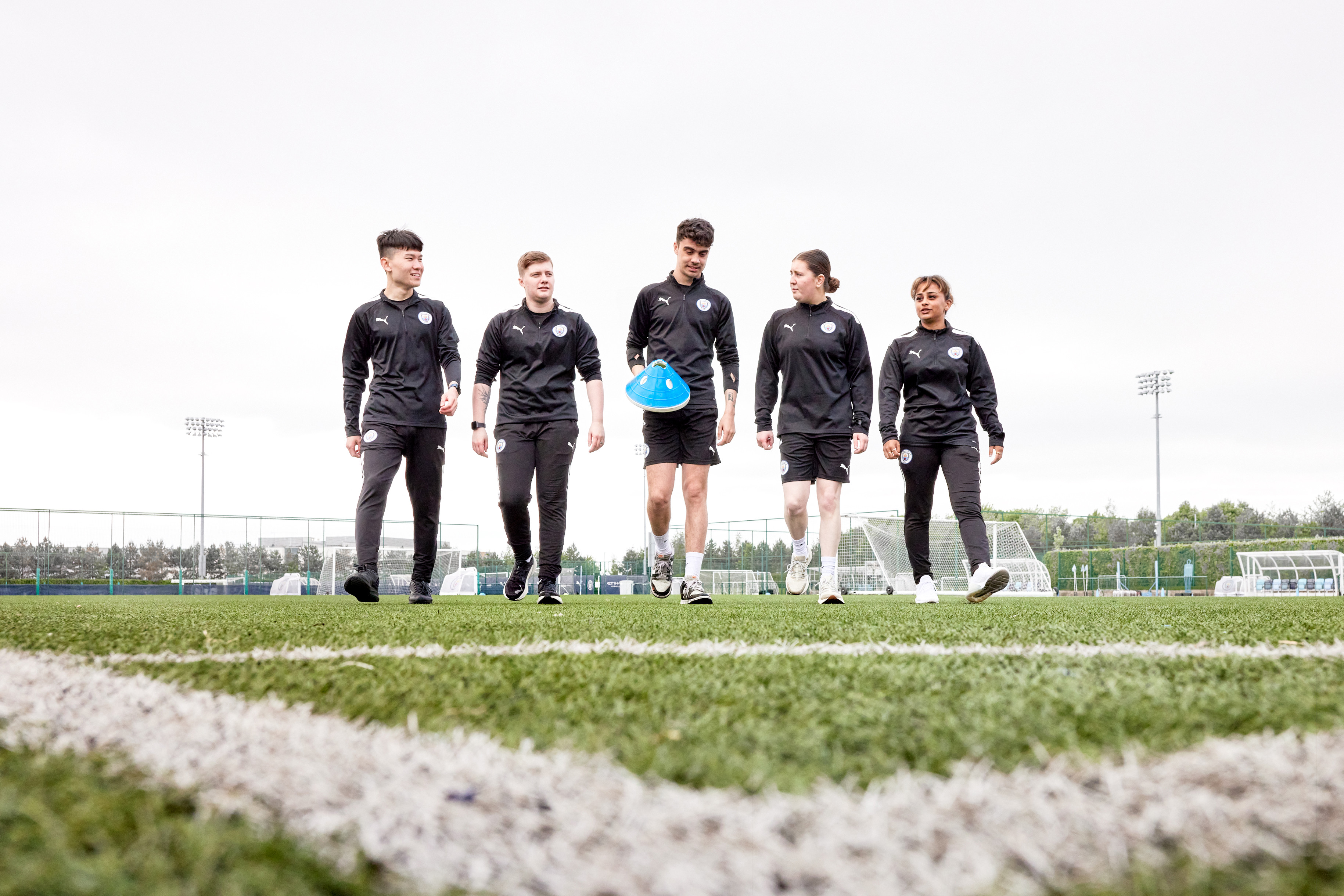Five individuals in Manchester City training attire walk confidently across a football pitch, one of them carrying a set of training cones.