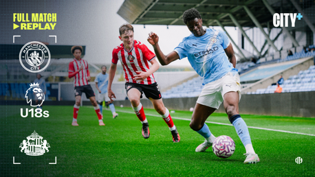 Manchester City U18 player in blue kit dribbling past a Sunderland U18 player in red and white kit during a match at the youth academy stadium.