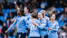 Group of Manchester City women's team players celebrating a goal on the field wearing blue jerseys.