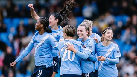 Group of Manchester City women's team players celebrating a goal on the field wearing blue jerseys.