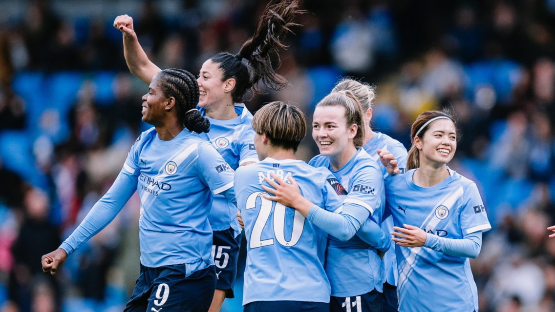 Group of Manchester City women's team players celebrating a goal on the field wearing blue jerseys.