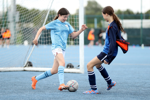 Two young girls playing soccer on an outdoor field, one in a light blue jersey dribbling the ball while the other in a dark blue jersey defends.