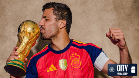 A soccer player wearing a Spain jersey kisses a replica of the FIFA World Cup trophy, celebrating with a fist pump. The image has a City at the World Cup watermark.