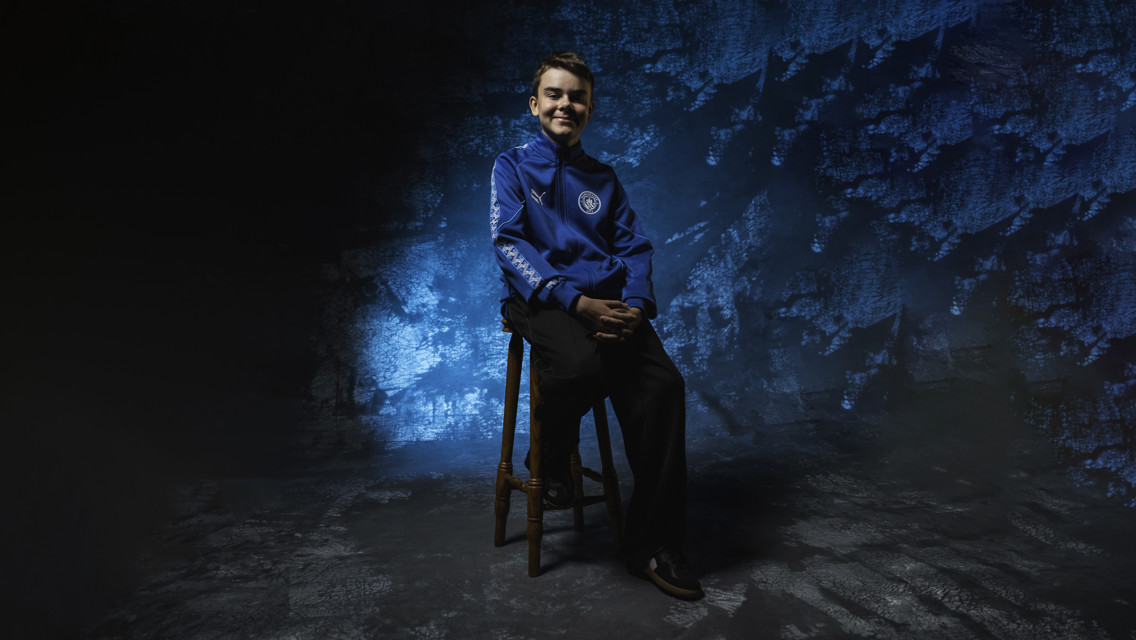 A young person wearing a blue Manchester City hoodie sits on a wooden stool against a textured blue background.