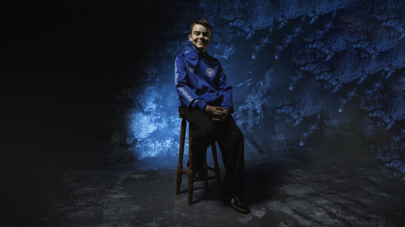 A young person wearing a blue Manchester City hoodie sits on a wooden stool against a textured blue background.