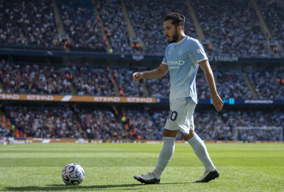 A Manchester City player in light blue kit walks on the pitch at a stadium, with a ball nearby, and a crowd in the background.