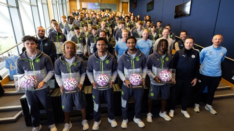 A group photo of Manchester City team members in a room, with some individuals holding footballs encased in display boxes. Manchester City apparel is worn by all team members.