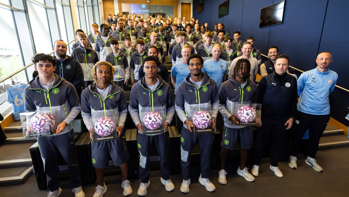 A group photo of Manchester City team members in a room, with some individuals holding footballs encased in display boxes. Manchester City apparel is worn by all team members.