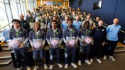 A group photo of Manchester City team members in a room, with some individuals holding footballs encased in display boxes. Manchester City apparel is worn by all team members.