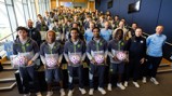 A group photo of Manchester City team members in a room, with some individuals holding footballs encased in display boxes. Manchester City apparel is worn by all team members.