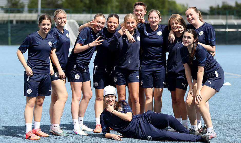 A group of players from Manchester City's Women's Team wearing blue training gear posing together on a football pitch. Most have their faces blurred.