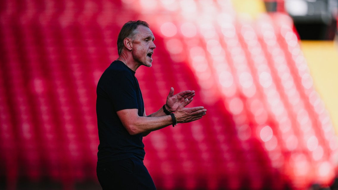 A soccer coach wearing a black shirt clapping and giving instructions from the sidelines in a stadium with red seating in the background.
