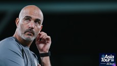 A bald man with a beard is wearing a gray shirt and a black watch, with his hand near his ear. The logo in the corner reads 'Asia Tour '26 presented by experience Abu Dhabi.'