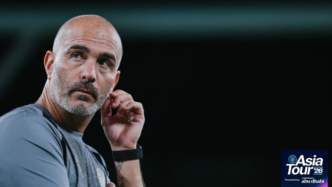 A bald man with a beard is wearing a gray shirt and a black watch, with his hand near his ear. The logo in the corner reads 'Asia Tour '26 presented by experience Abu Dhabi.'
