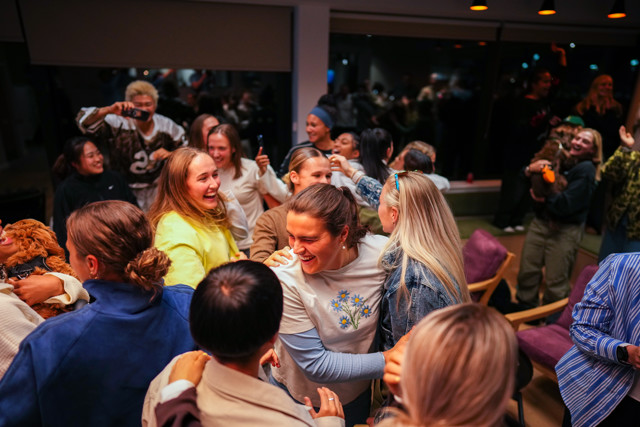 A group of people gathered indoors, engaging in a lively celebration. Some individuals hold drinks, and a person in the background is holding a dog.